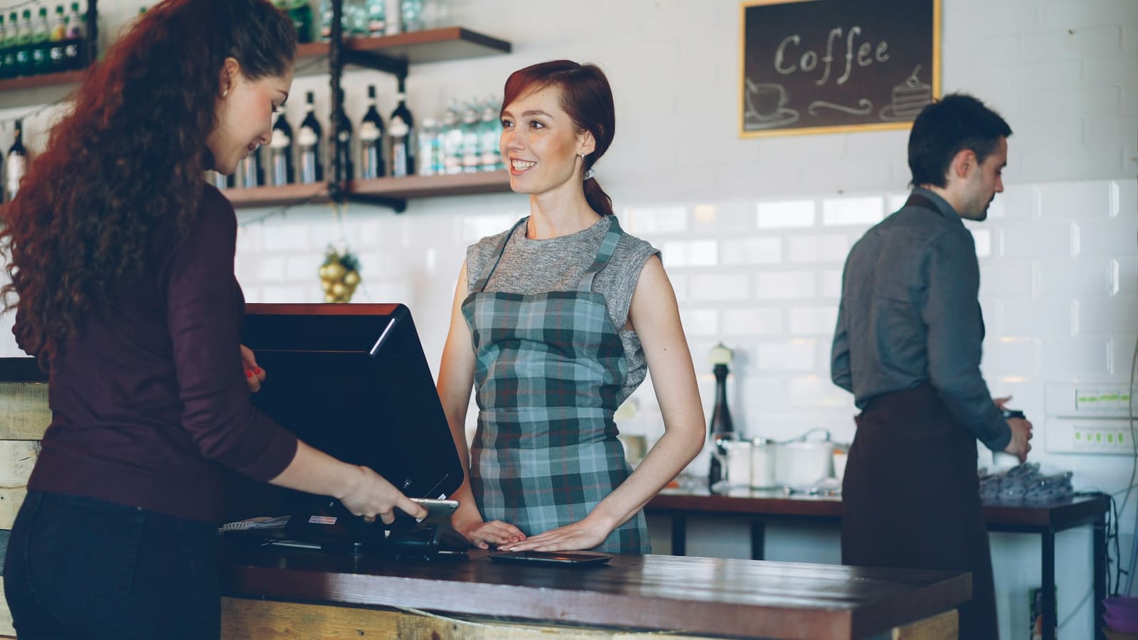 Una clienta paga con el teléfono en el mostrador de una cafetería mientras la barista sonríe.